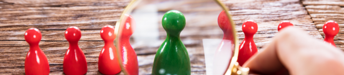 grüne Spielfigur inmitten von roten wird durch eine Lupe vergrößert